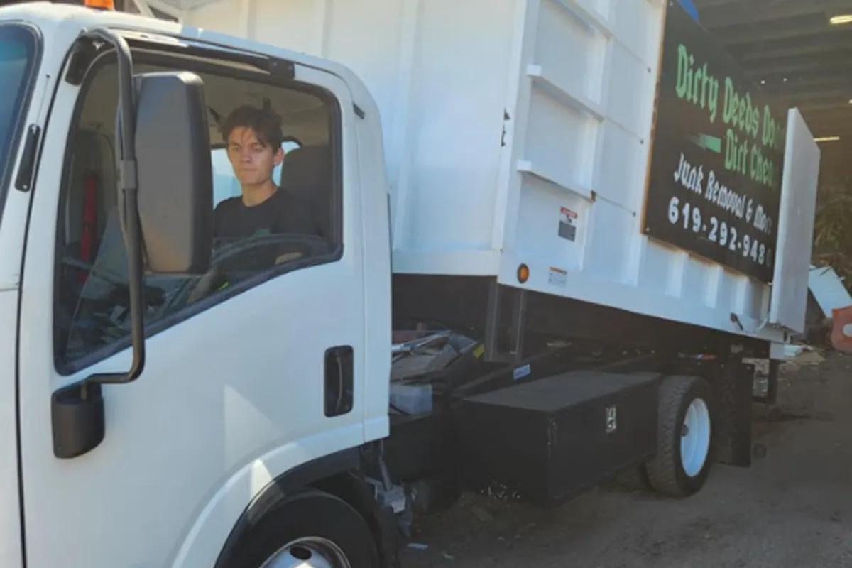 Junk removal truck with company branding parked in a residential alley, featuring visible signage reading “Dirty Deeds Done Dirt Cheap Junk Removal & More 619-292-5488,” with a young driver seated in the cab; white hauling vehicle used for waste disposal, property cleanouts, and debris removal services, emphasizing professional junk hauling business operations.