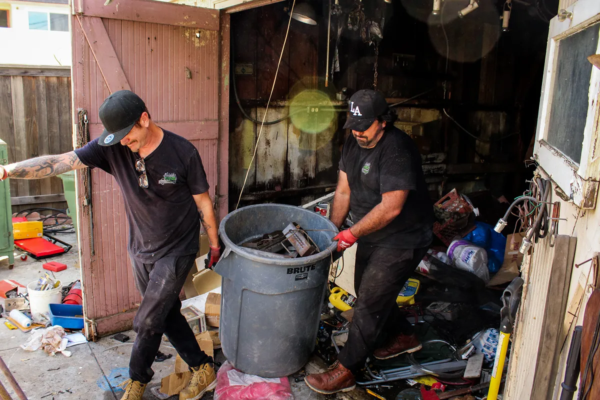 Two junk removal workers clearing out a cluttered garage, pushing a large BRUTE trash bin filled with scrap materials and debris; both men wear black work shirts, gloves, and caps, surrounded by scattered tools, boxes, and household junk in a messy residential cleanup scene, illustrating professional hauling, garage cleanout, and waste disposal services.