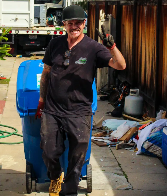 Smiling junk removal worker in black cap and gloves flashing a peace sign in a residential alley, standing near a blue recycling bin, propane tank, and cleanup equipment with a junk removal truck in the background; casual laborer with visible arm tattoos representing professional hauling, waste disposal, and property cleanout service.
