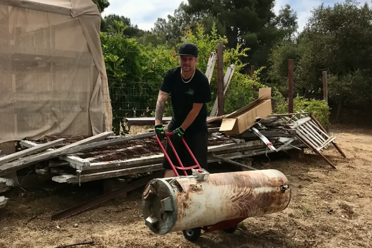 Junk removal worker using a hand truck to transport a large rusty cylindrical tank in an outdoor yard, surrounded by stacked scrap wood, metal debris, and discarded materials; man wearing black work clothes, gloves, and cap performing heavy lifting and property cleanup, illustrating professional hauling, bulk item removal, and outdoor junk disposal services in a rural or residential setting.