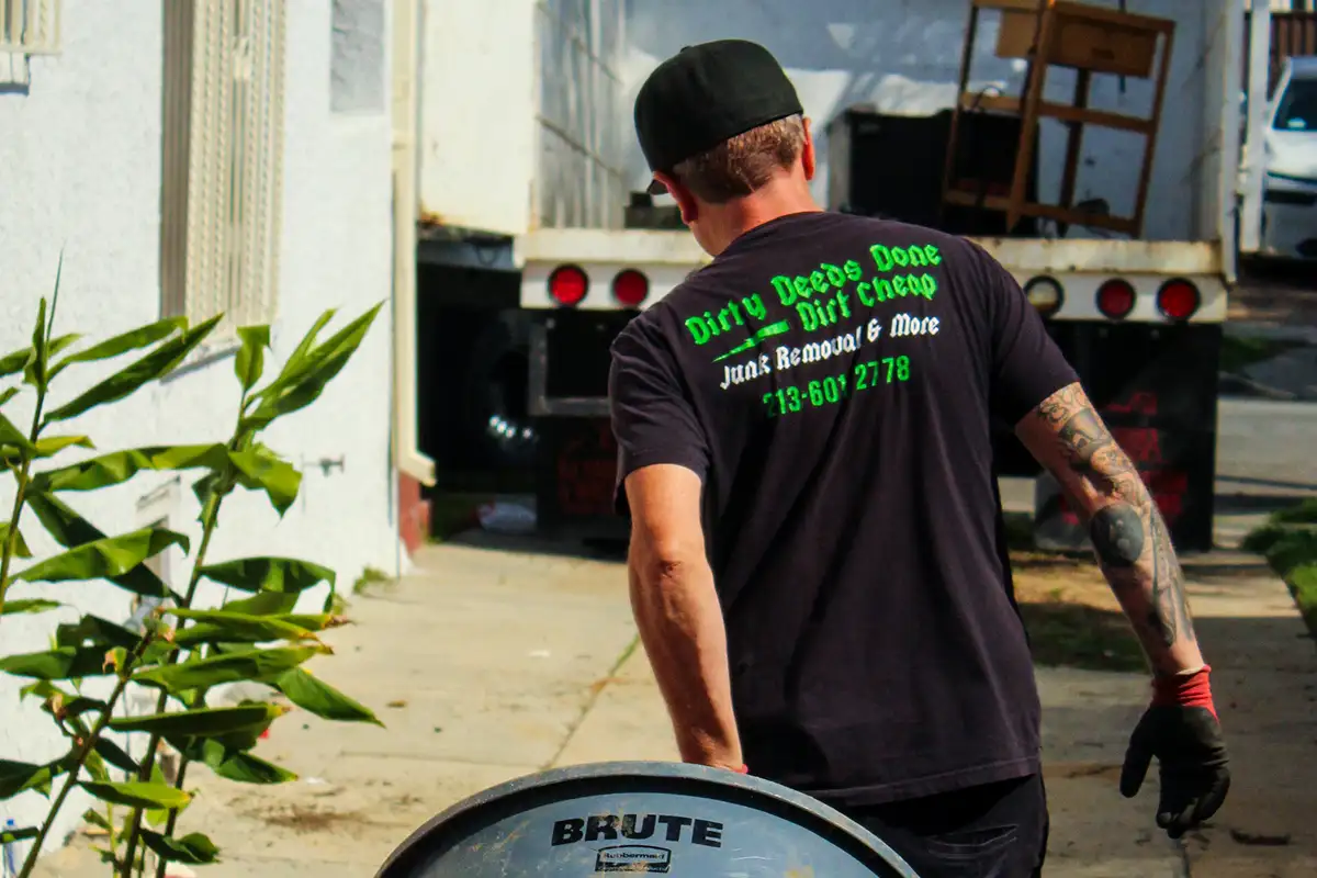 Man in black T-shirt performing junk removal work, carrying a large BRUTE trash bin toward a truck in a residential alley, wearing gloves and cap, visible arm tattoos; shirt text reads “Dirty Deeds Done Dirt Cheap Junk Removal & More 713-601-2778,” conveying professional hauling, waste disposal, and cleanup service in an outdoor urban setting with greenery and driveway background.