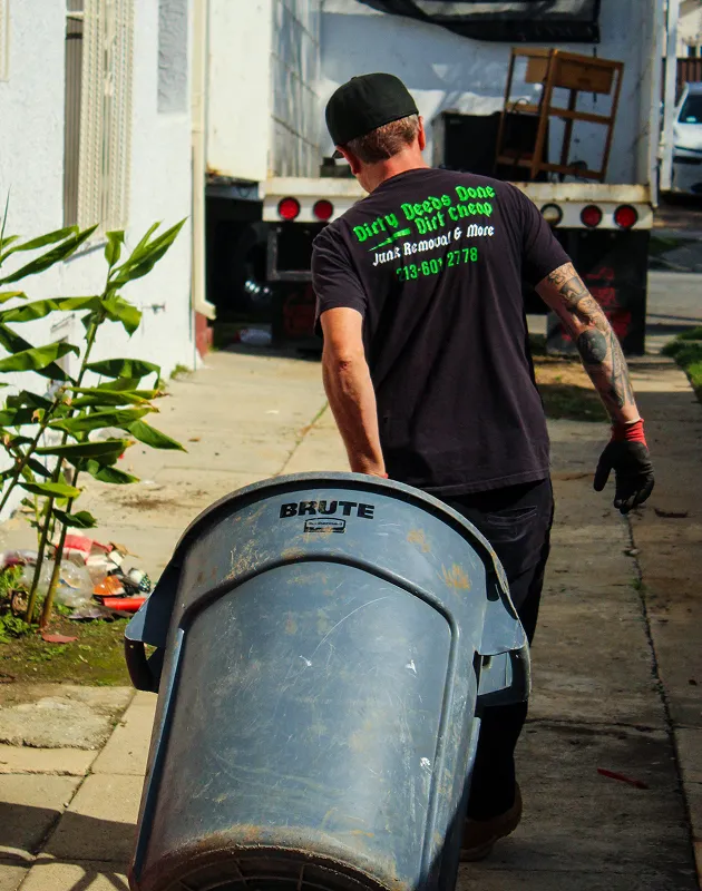 Man in black T-shirt performing junk removal work, carrying a large BRUTE trash bin toward a truck in a residential alley, wearing gloves and cap, visible arm tattoos; shirt text reads “Dirty Deeds Done Dirt Cheap Junk Removal & More 713-601-2778,” conveying professional hauling, waste disposal, and cleanup service in an outdoor urban setting with greenery and driveway background.